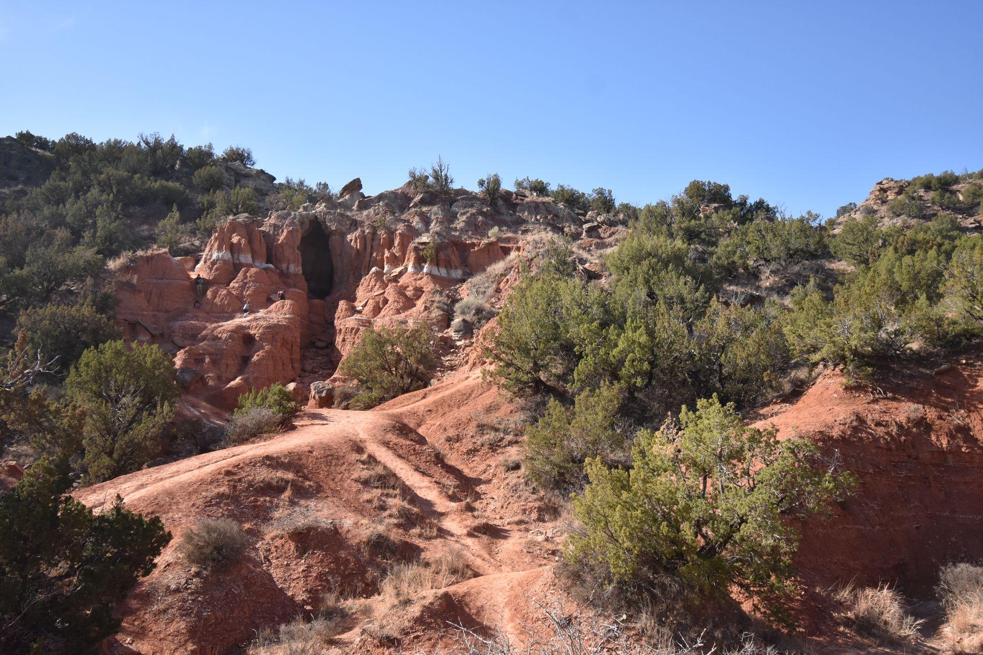 A path leading up to a orange cave in the background. Around the cave is a stripe of white in the orange rocks.