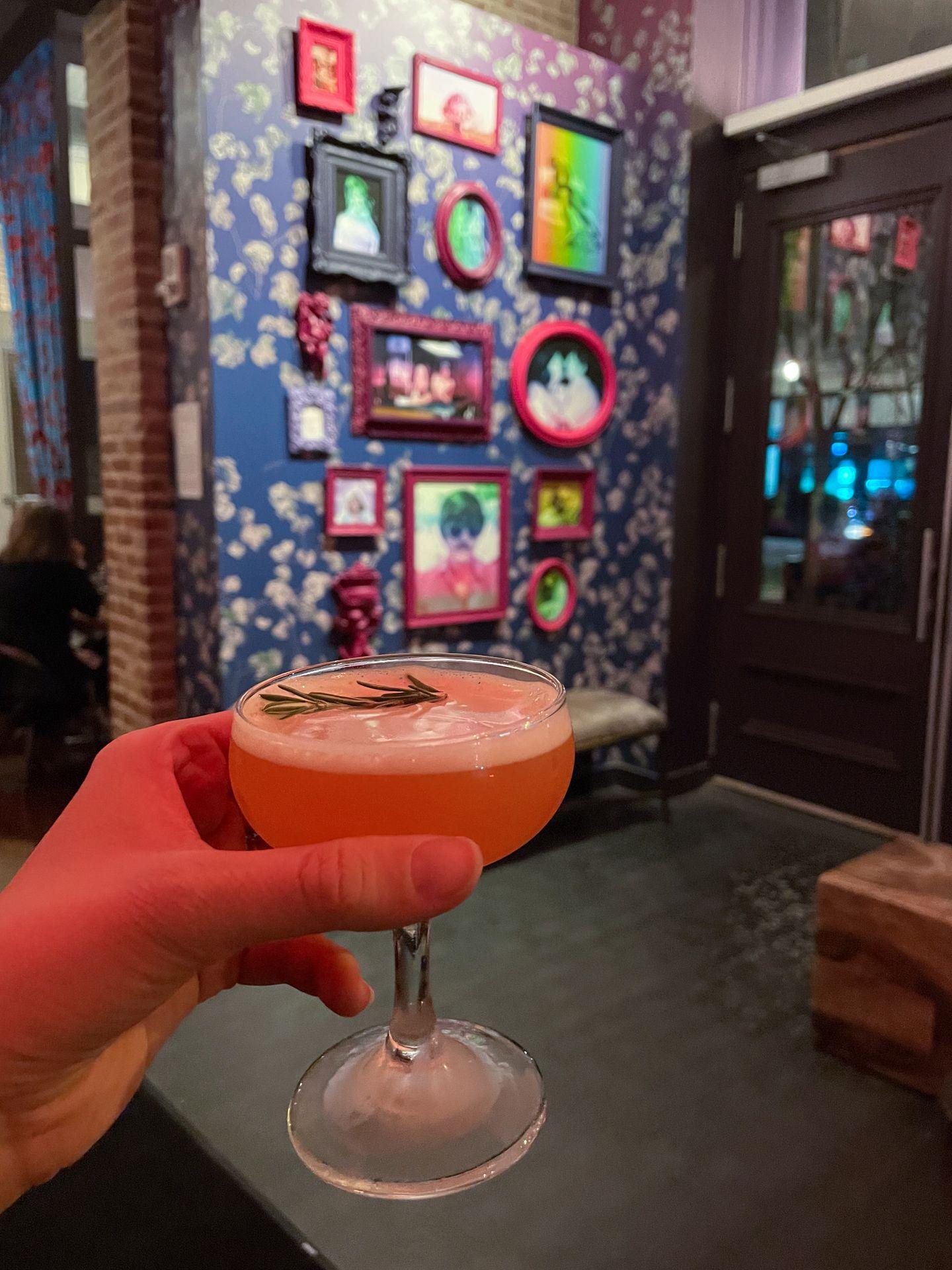 Holding up a cocktail in a room with a vibrant wall full of artwork