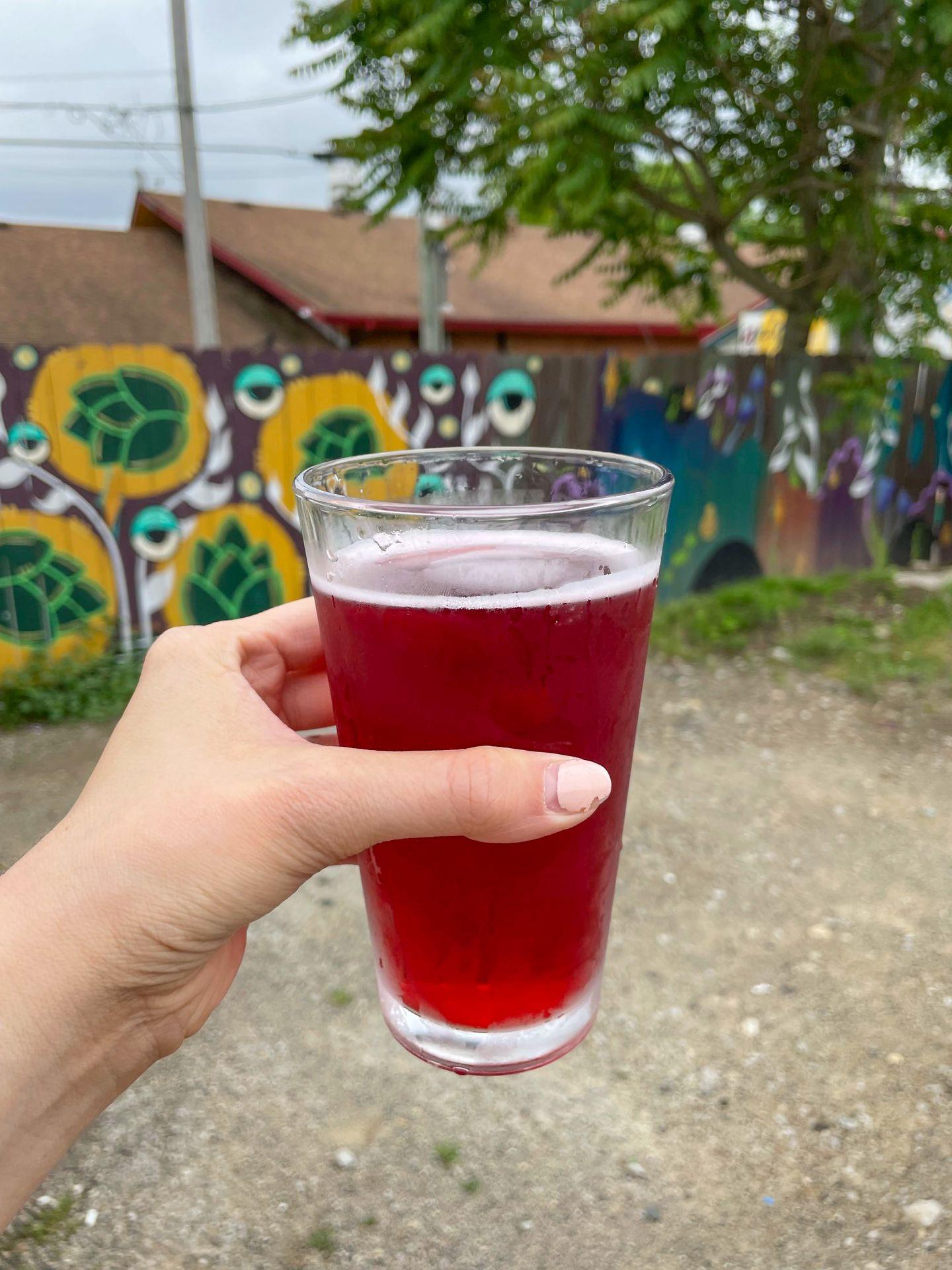 Holding up a beer on the patio of Trail Town Brewing. The background is a colorful fence painted with murals