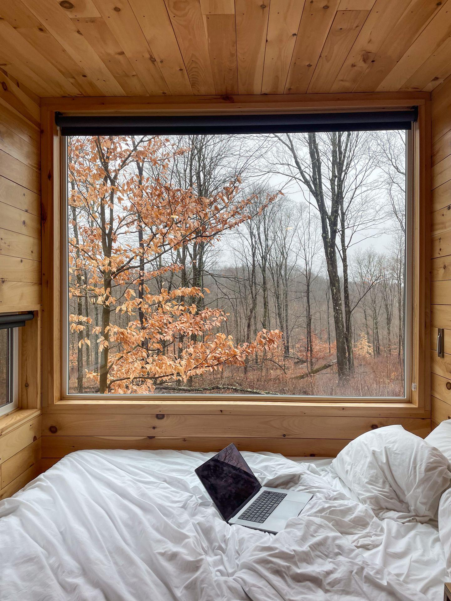 Looking out a large window inside of a Postcard Cabin. A laptop sits on the bed