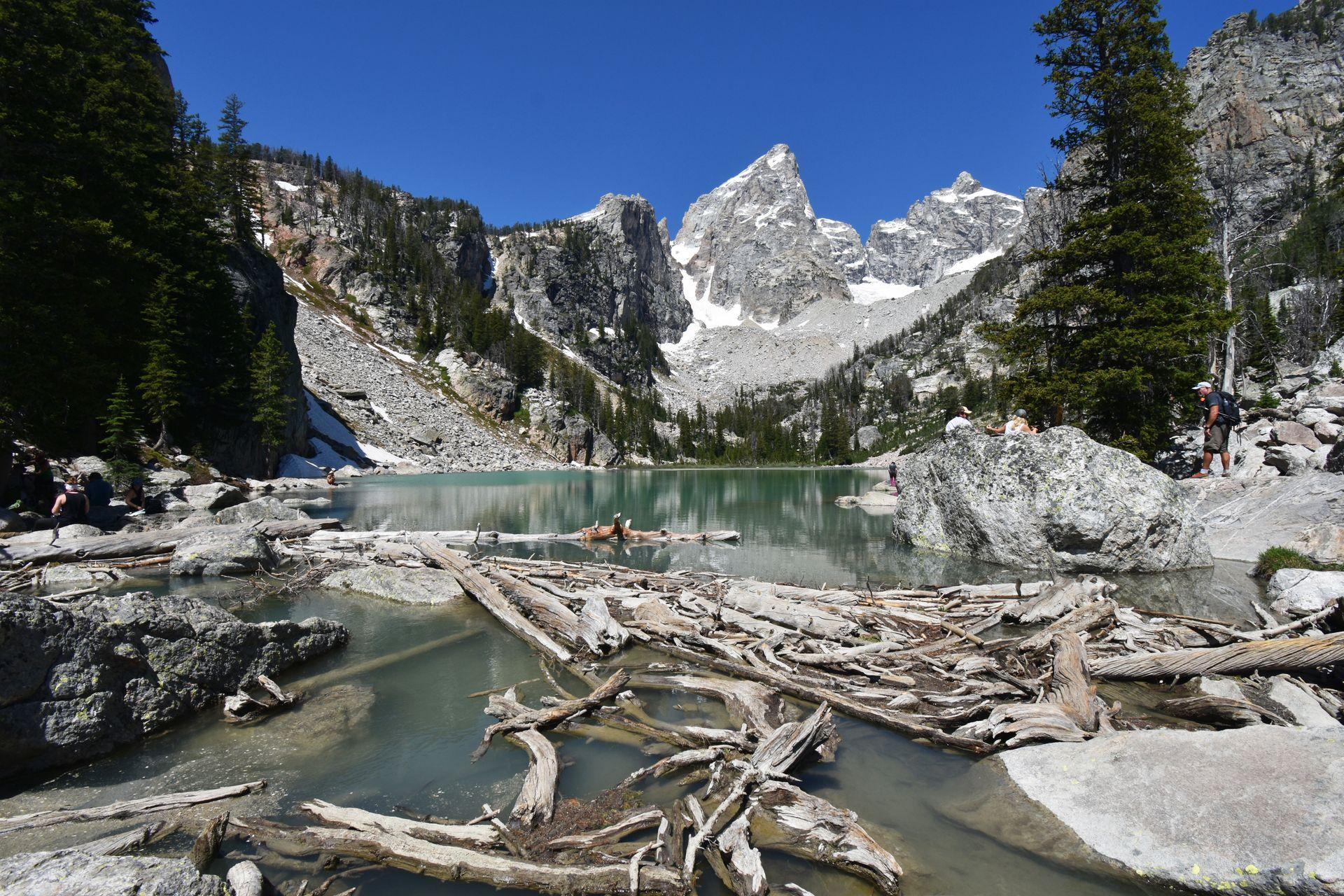 An aqua blue lake with pointed mountains in the distance. There are a couple large boulders in the lake.