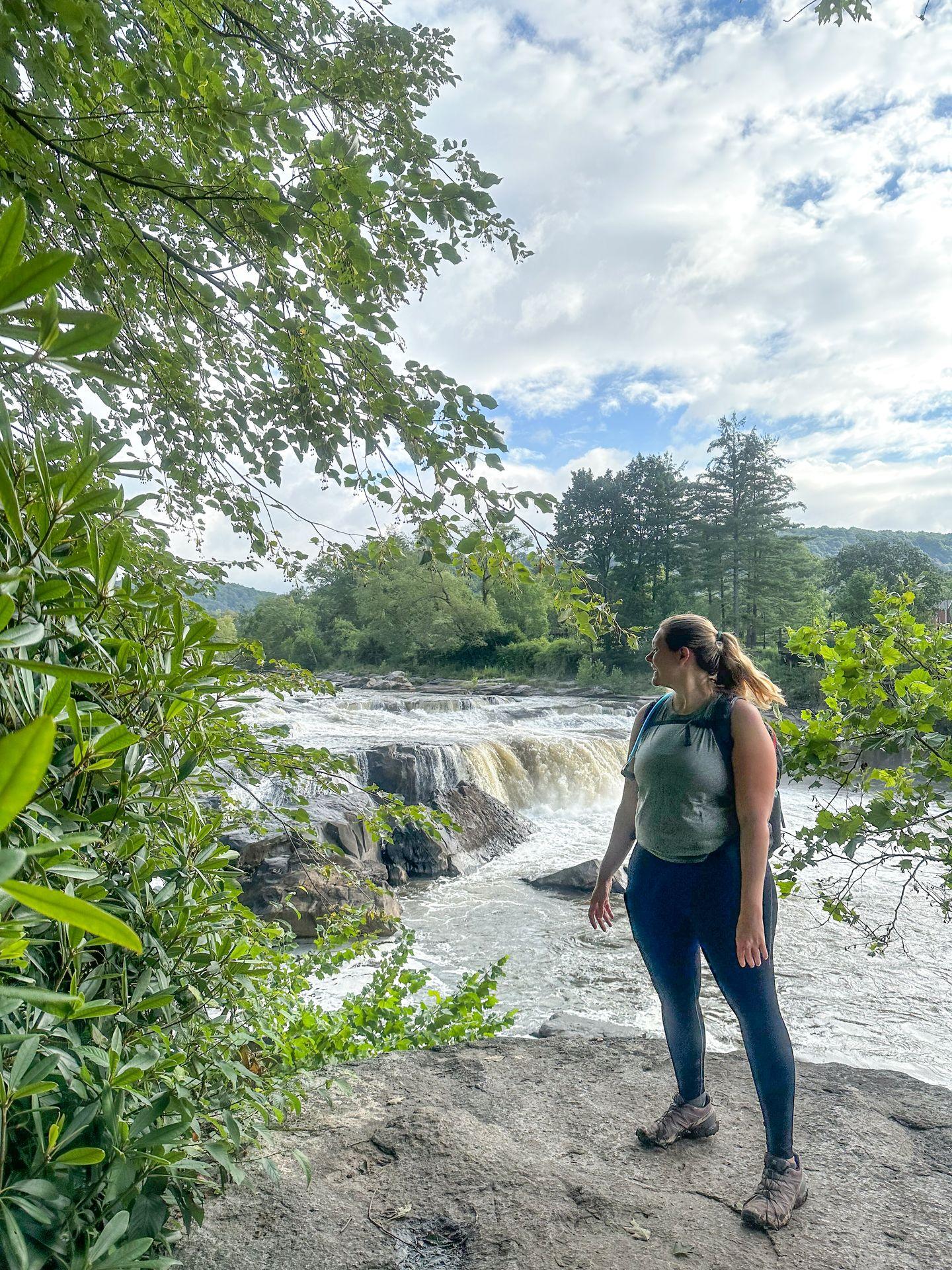 Lydia standing to the right with a waterfall in the background along the Ferncliff Trail in Ohiopyle State Park.