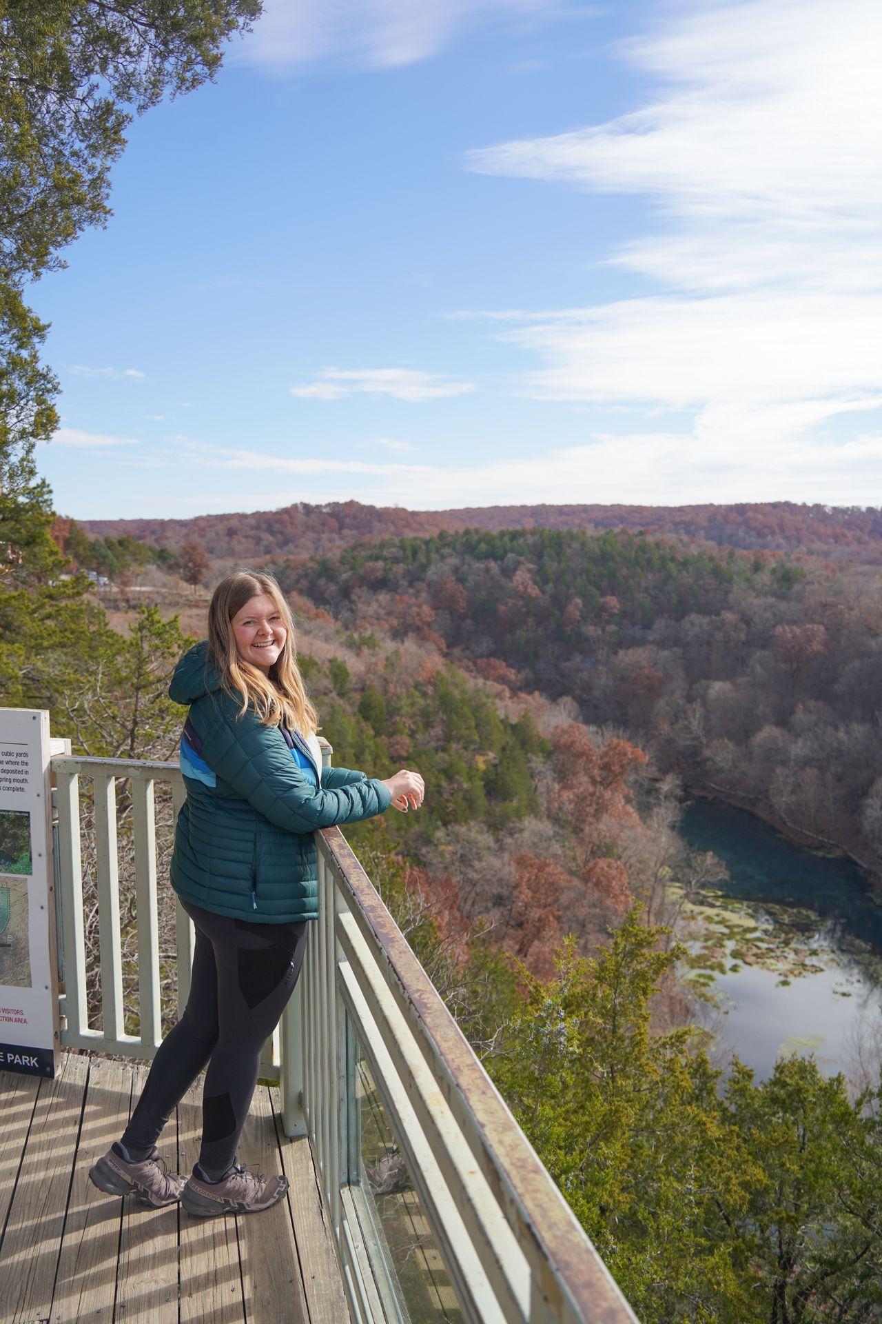 Lydia standing at an overlook high above a river. There is some fall foliage on the trees