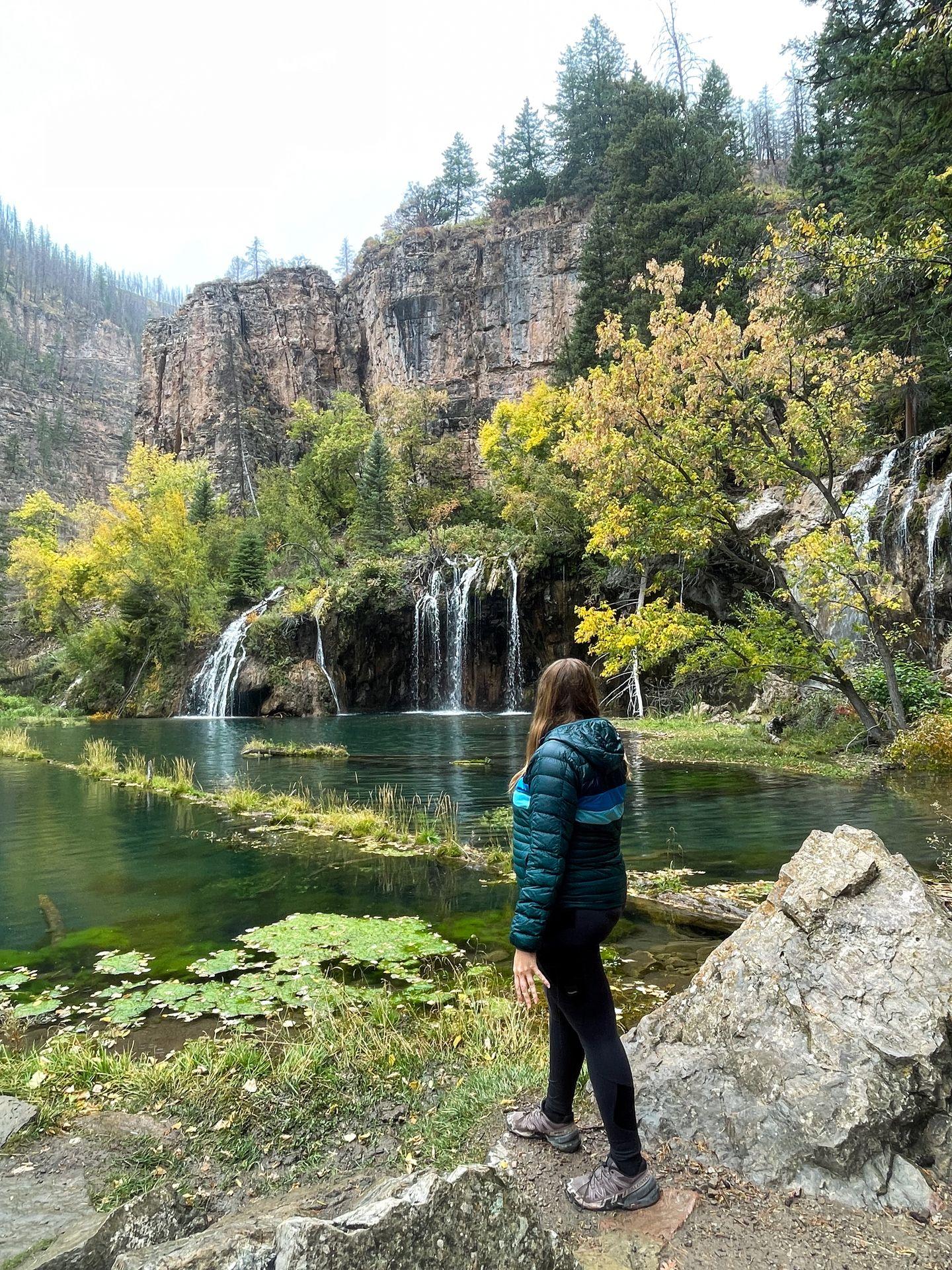 Lydia looking at a green lake with a wide waterfall flowing into it. There is also a tree with yellow foliage.