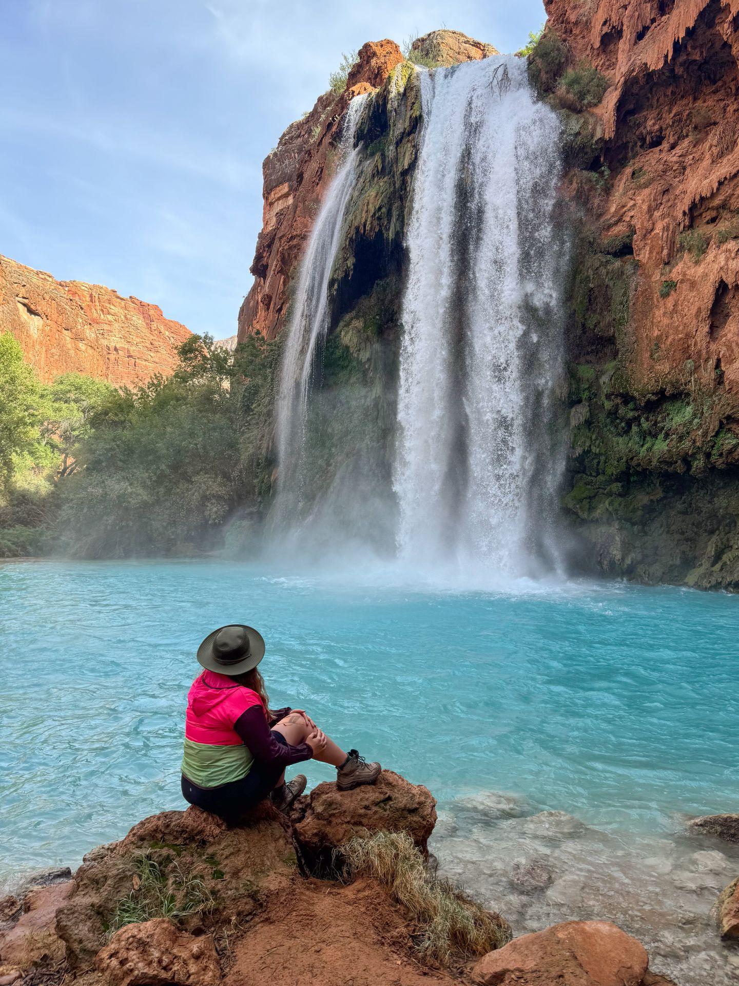 Lydia sitting and looking up at Havasu Falls, which is falling into a pool of aqua-blue water