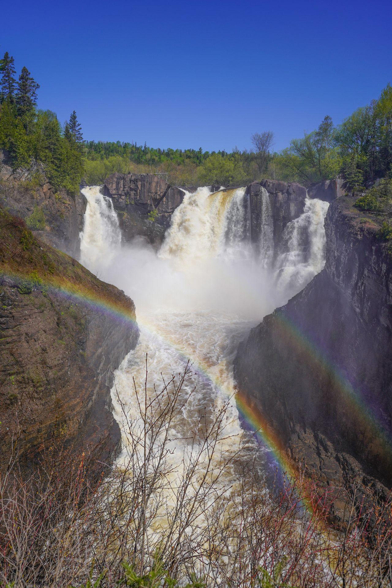 A waterfall flowing down into a black gorge from 3 directions. A rainbow is in the foreground.