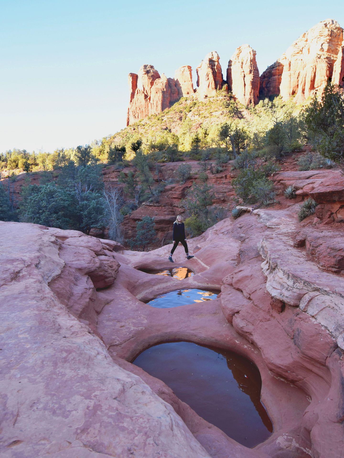 Lydia walking behind 3 pools of water. There are jagged, orange rocks in the background.