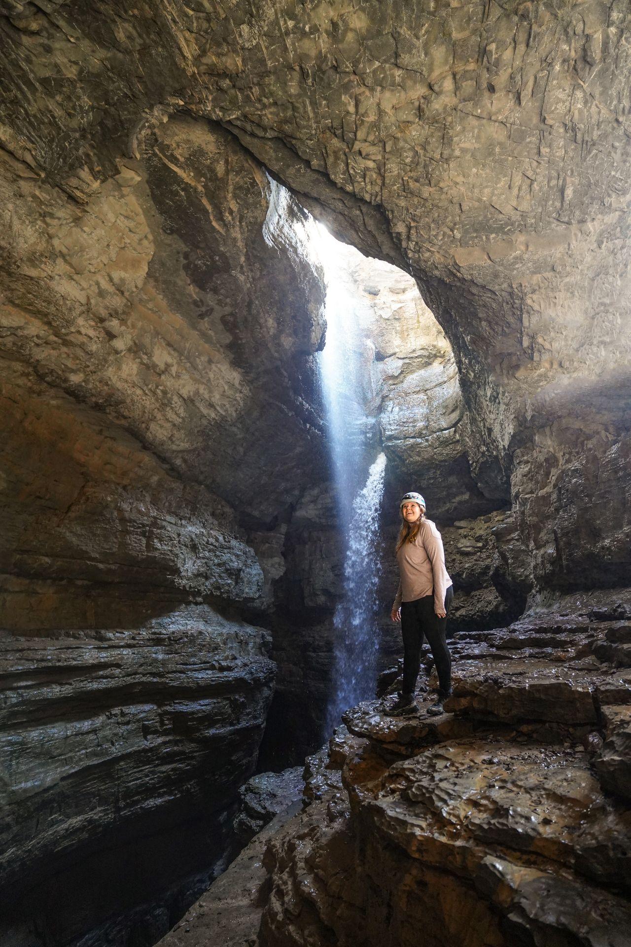 Lydia in a cave and wearing a helmet, next to a waterfall coming down through a hole in the top of the cave