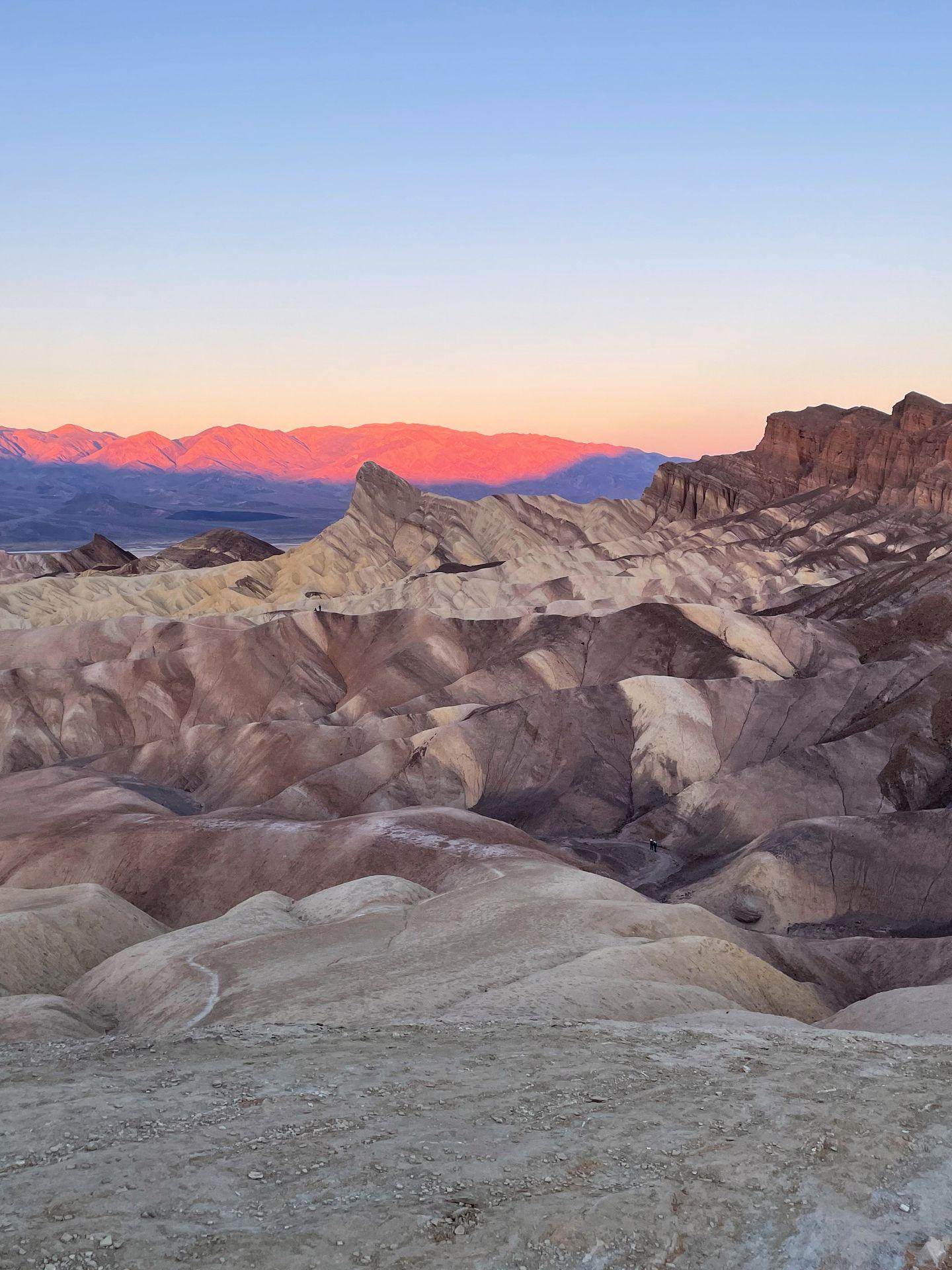 A scene with badlands that look to be striped in various shades of brown. The mountains in the distance glow pink and blue from the rising sun.