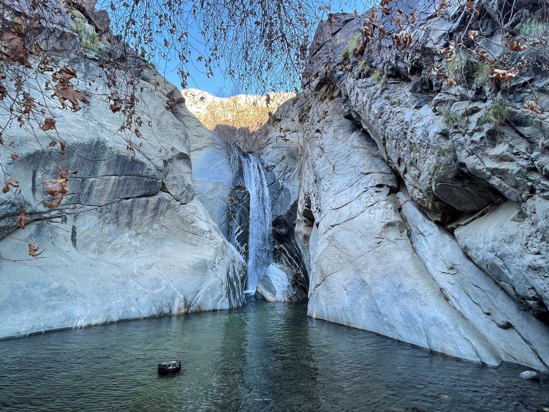 A waterfall flowing into a pool of green water. The rock face next to the waterfall has some interesting abstract white stripes.