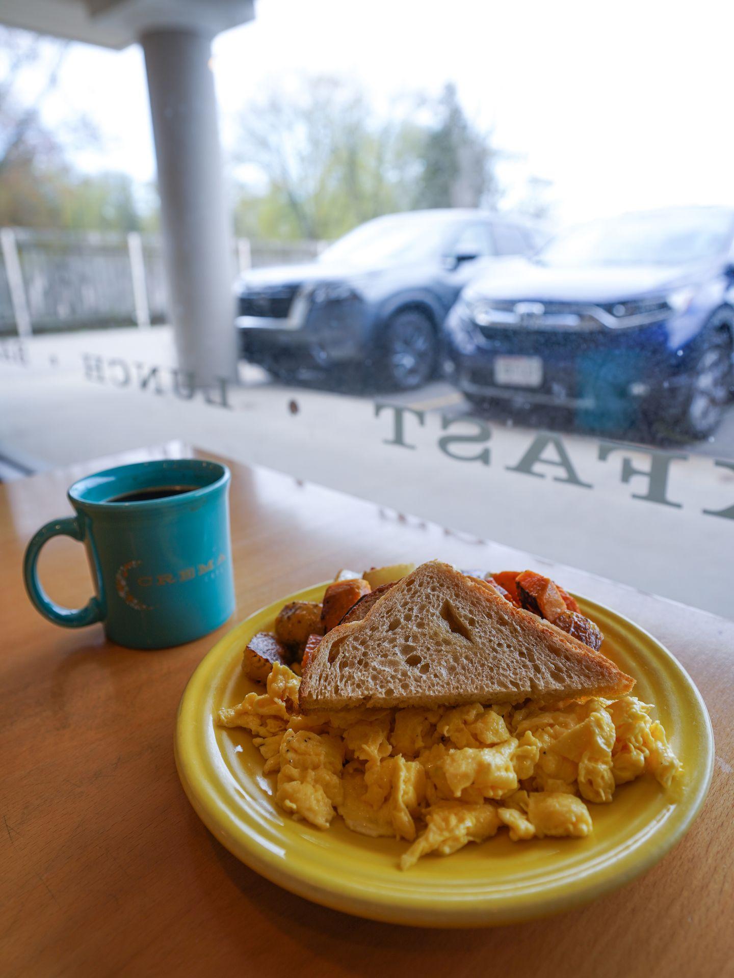 A yellow plate with scrambled eggs, toast and sweet potato has. In the background, there is a window and a blue mug with coffee.