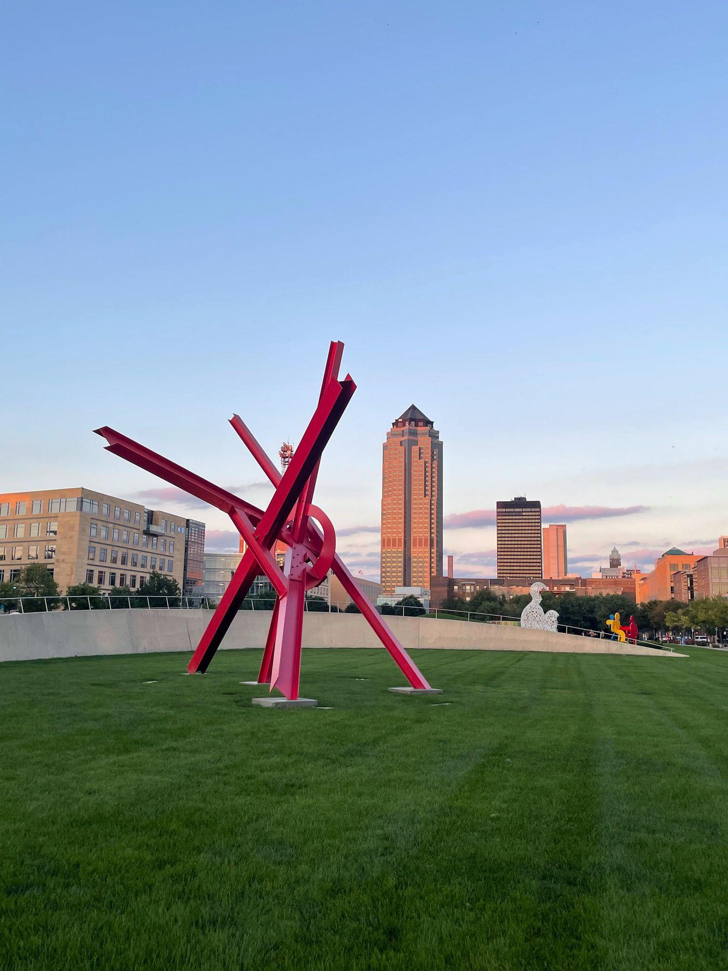 A red sculpture in a lawn, with tall buildings in the distance, in downtown Des Moines