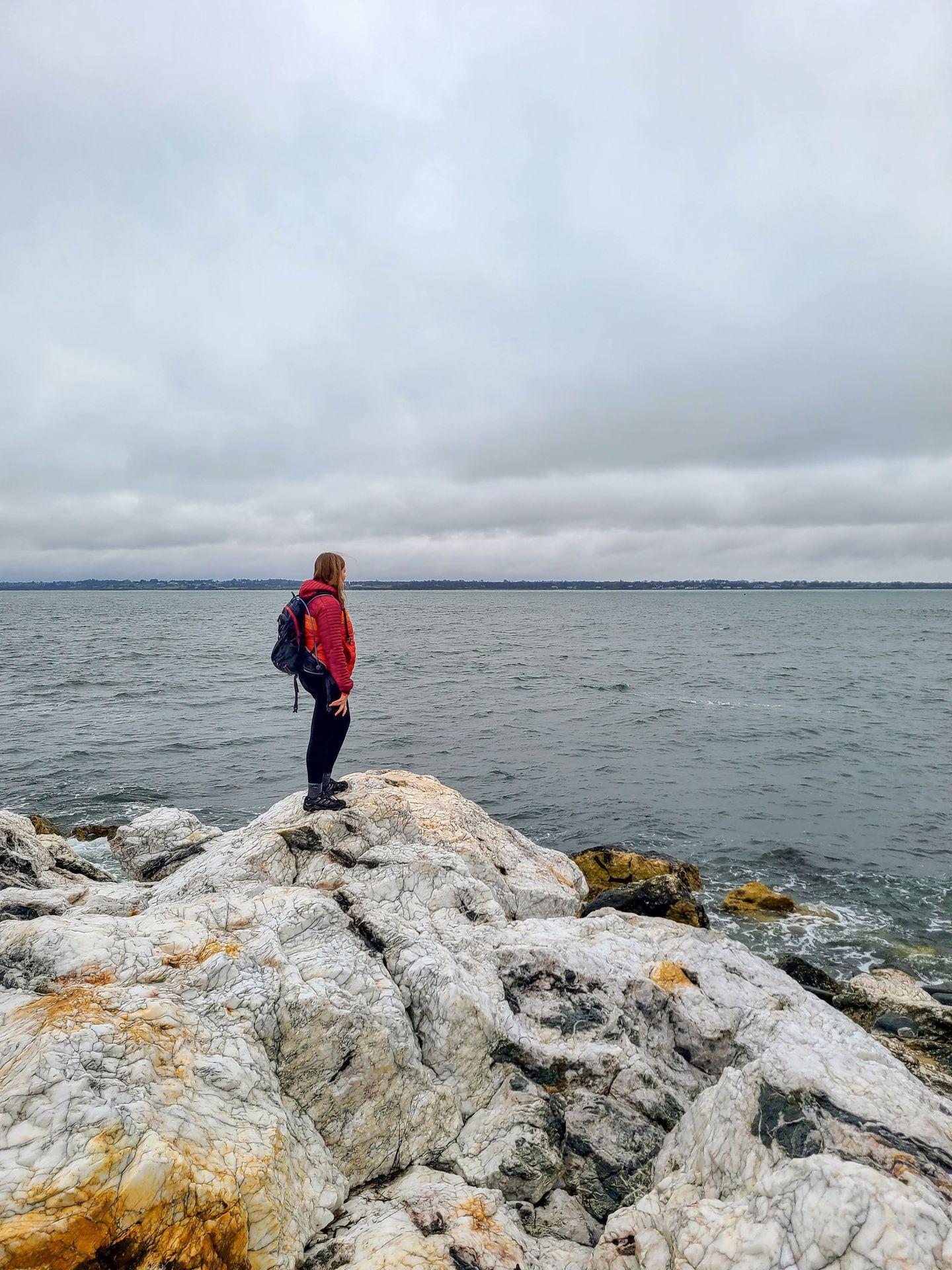 Lydia standing on top of a white rocks with a few colorful areas of orange. She is looking out at the ocean.