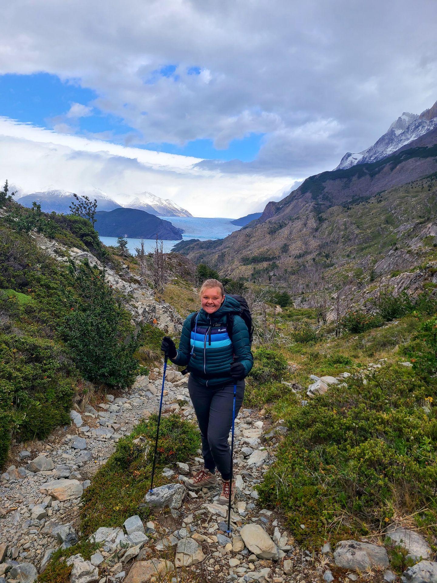 Lydia on a trail with a glacier in the background. She is wearing a Cotopaxi puffer jacket and holding hiking poles.
