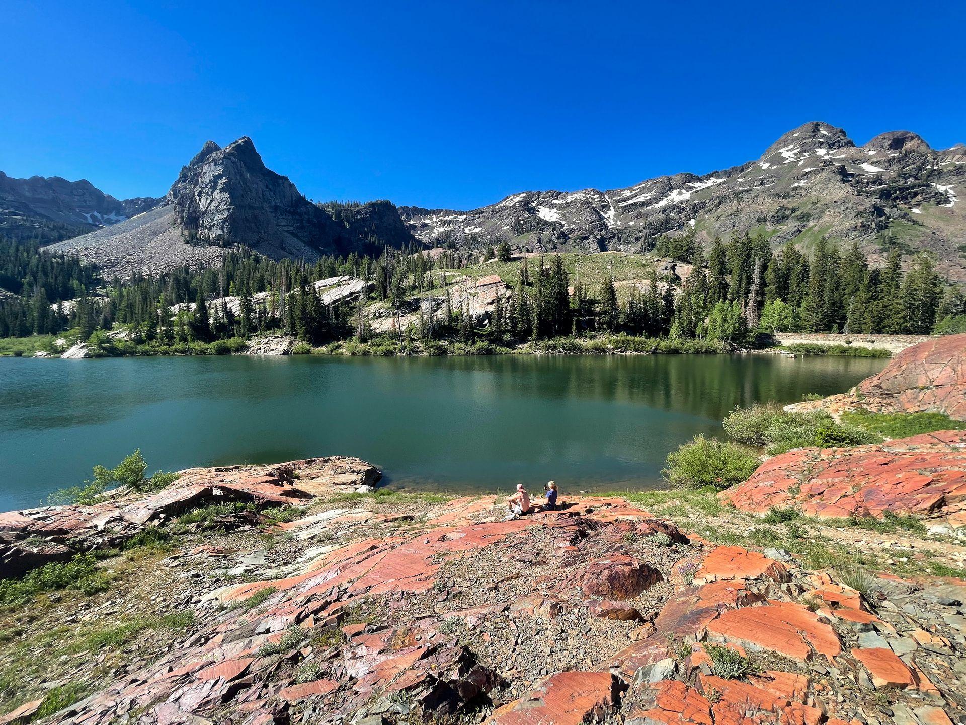 A view of Lake Blanche. Two hikers sit on some red rocks near the lake and a pointy mountain peak is seen on the opposite side of the water.