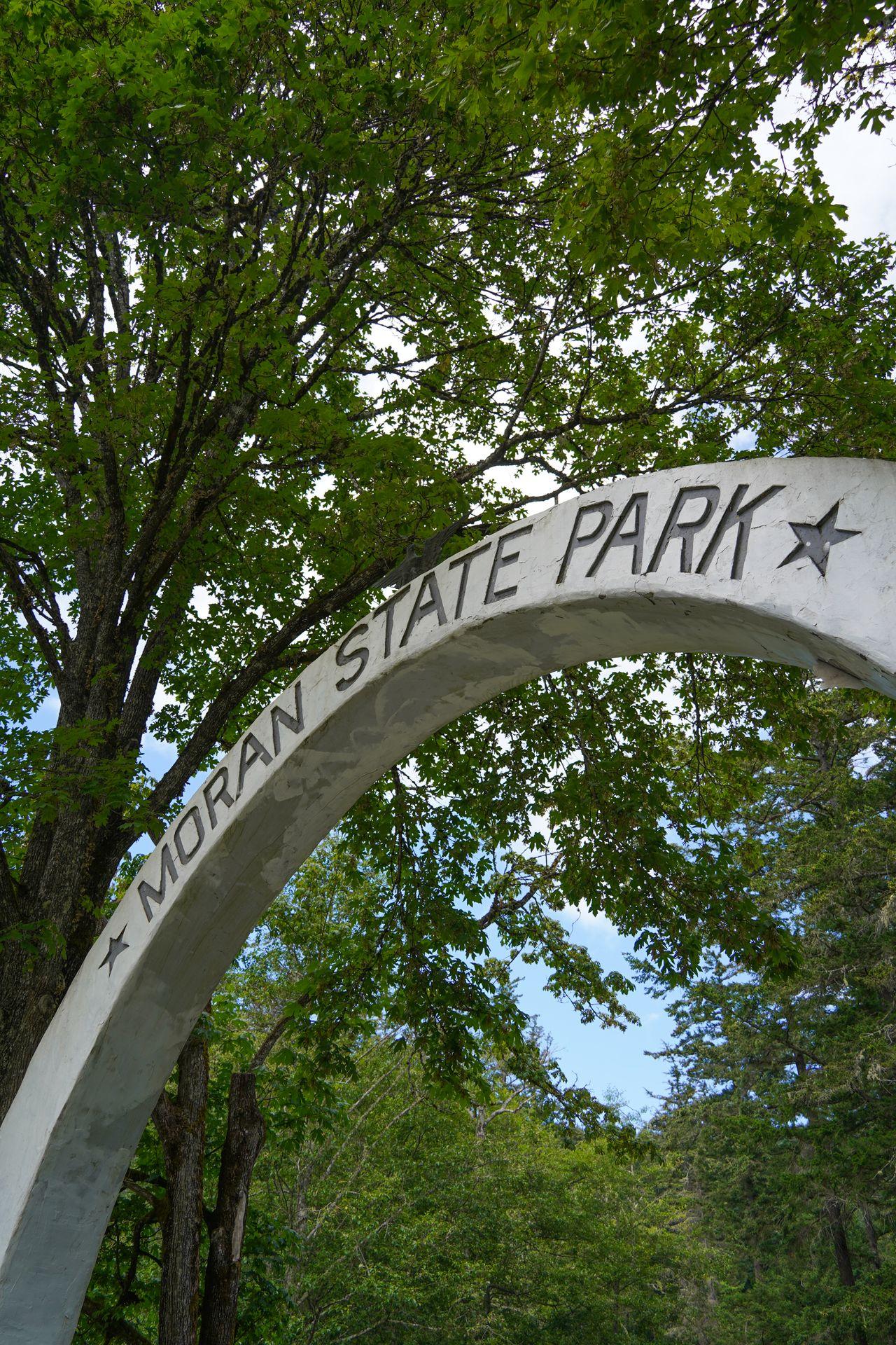 Looking up at a white arch that reads 'Moran State Park.' It extends over the road as you enter the park