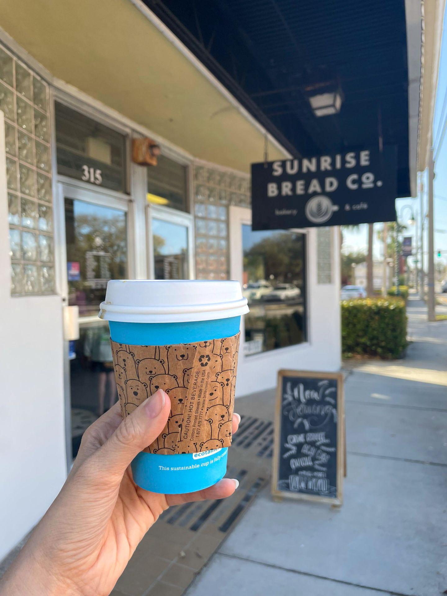 Holding up a blue, paper coffee mug in front of Sunrise Bread Co.