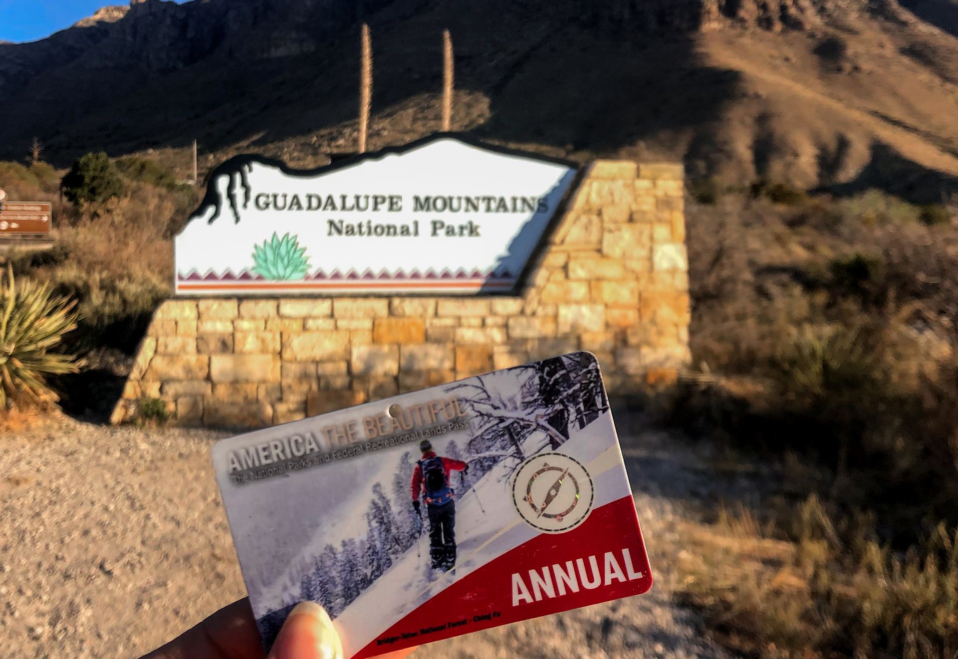 Holding up an America the Beautiful national park pass in front of an entrance sign at Guadalupe Mountains