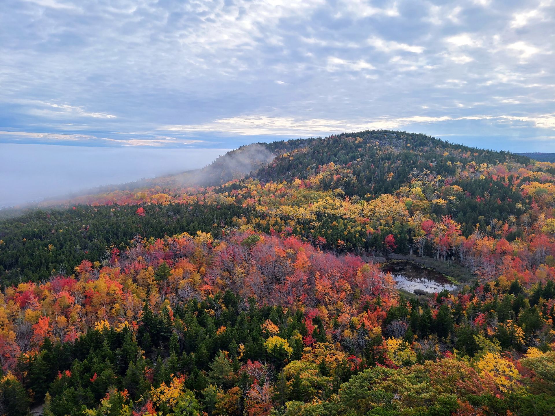 A view from the top of the Beehive Trail. There are rolling hills covered in green trees along with red, orange and yellow foliage. There is a small lake nestled among the trees.