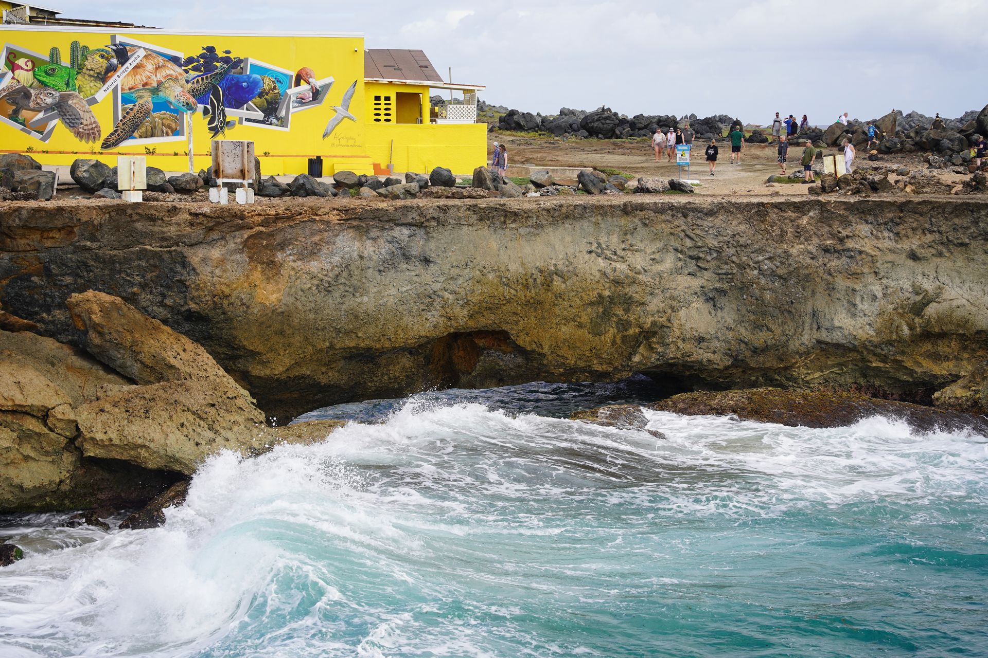 A wave crashing up below the natural bridge. A bright yellow building is on land above the bridge