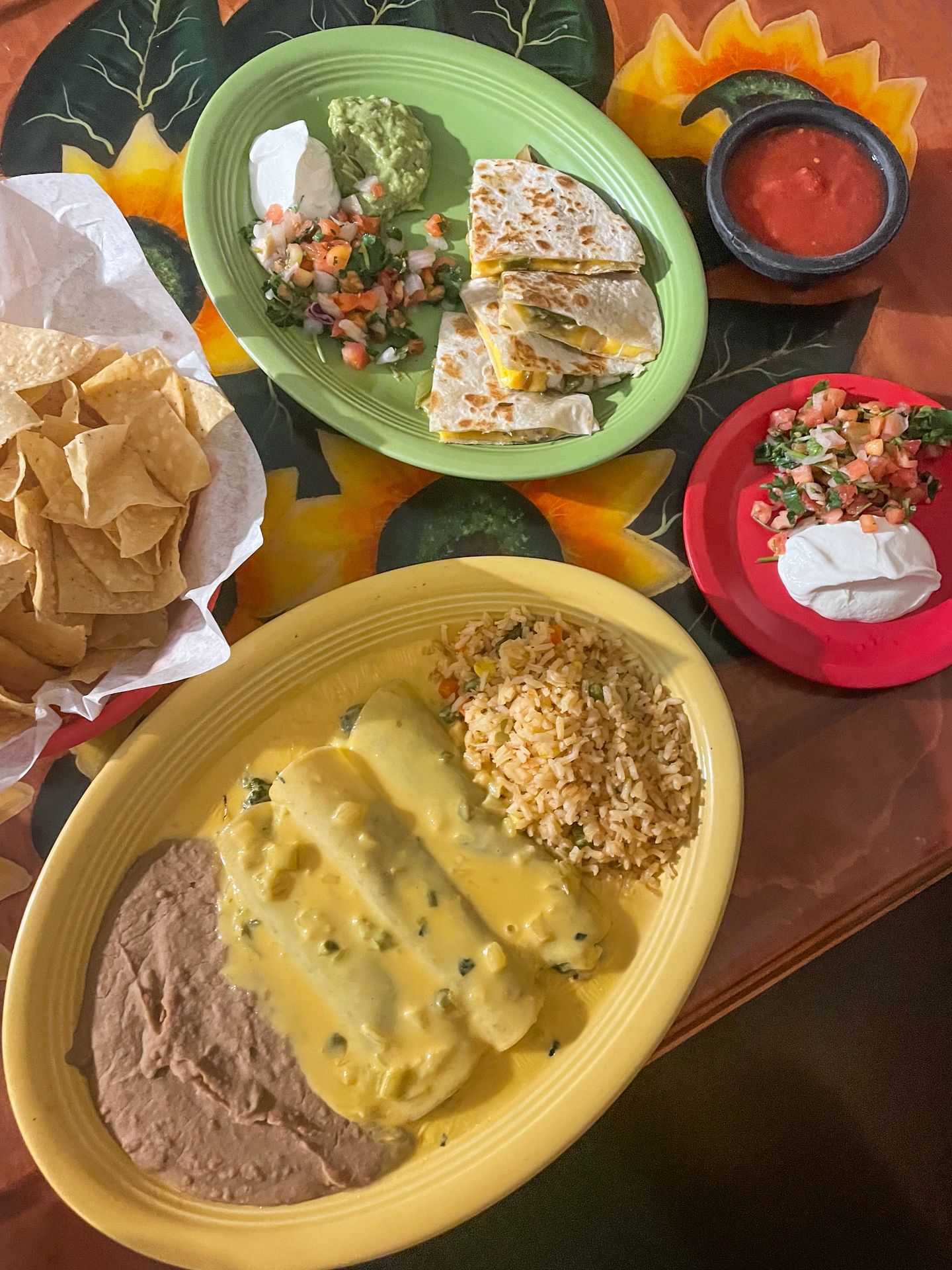 A couple colorful plates of Mexican food, next to a bowl of chips, at Carmela's. There are enchilada's served with rice and beans and quesadillas served with guacamole, sour cream and pico de galo.