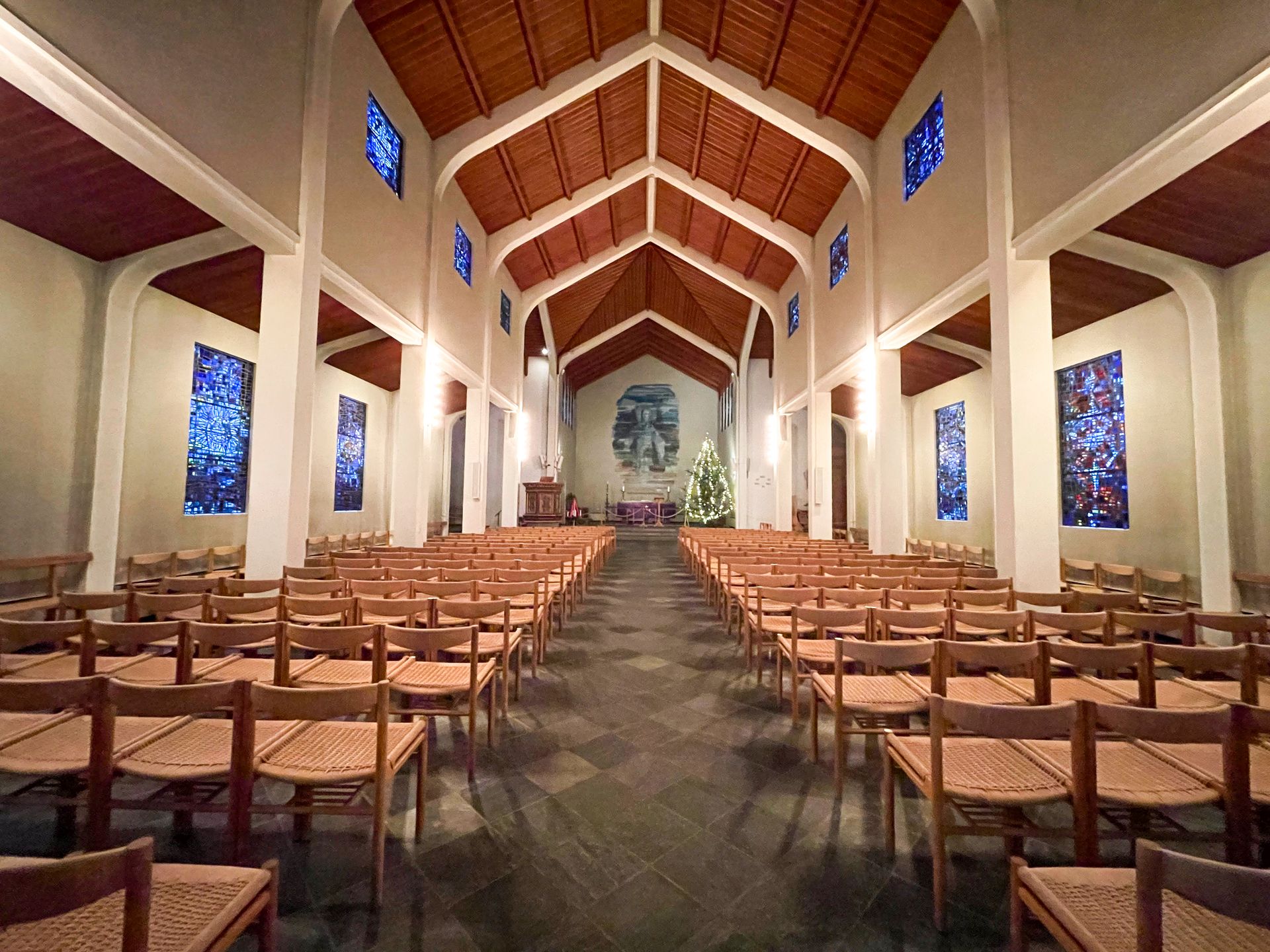 The interior of Skalholt Church. The walls are white with a brown ceiling and brown chairs. There are stained glass windows on the walls.