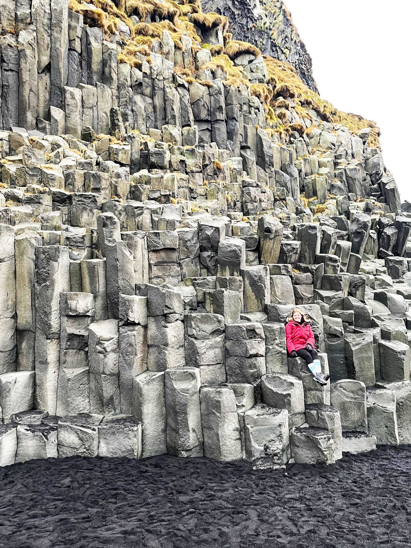 Lydia sitting on a wall of column-shaped rocks at Reynisfjara Beach.