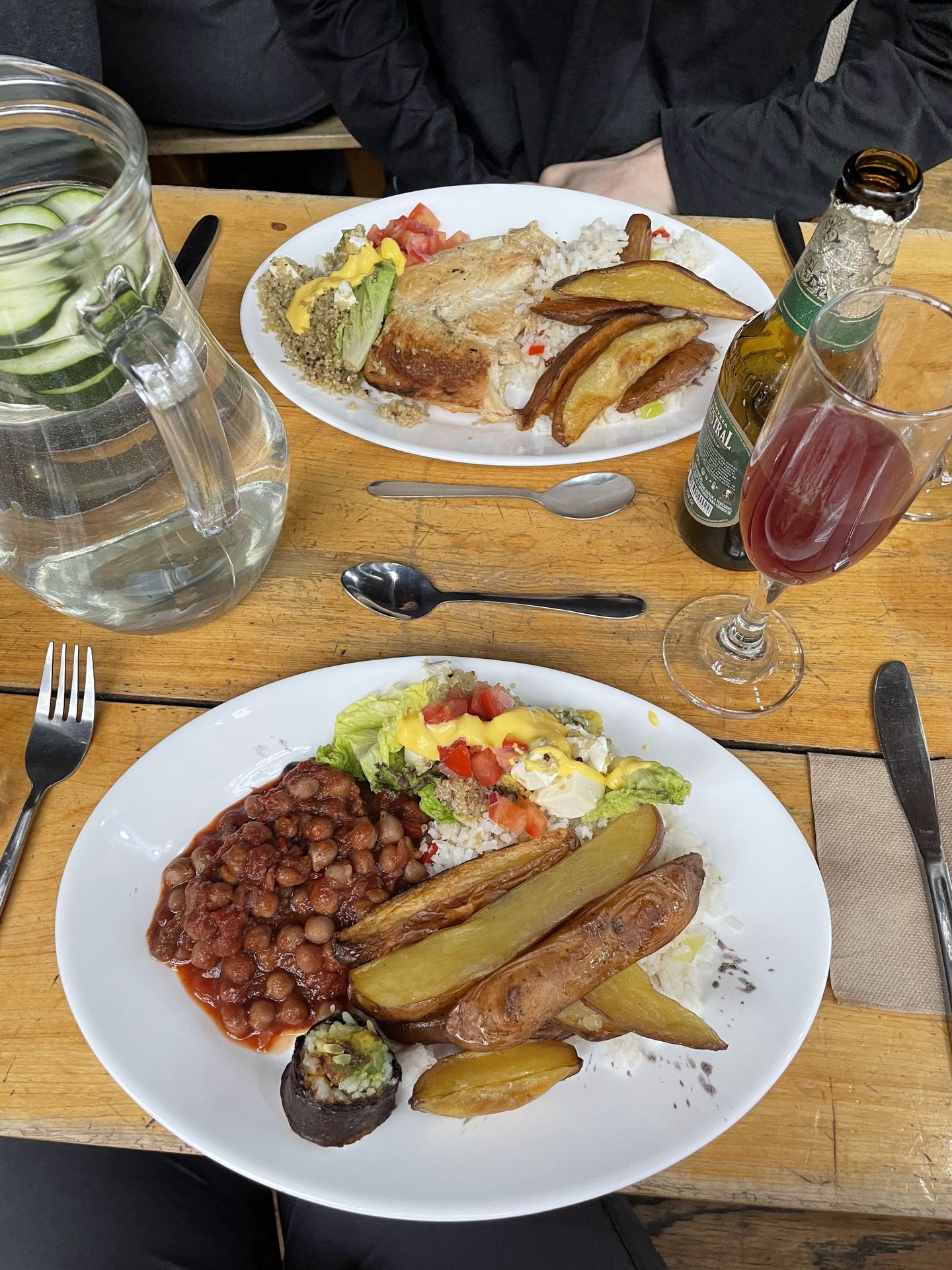 A plate of food that includes potato wedges, salad, baked beans and a sushi roll. There is a Calafate Sour cocktail next to the meal.