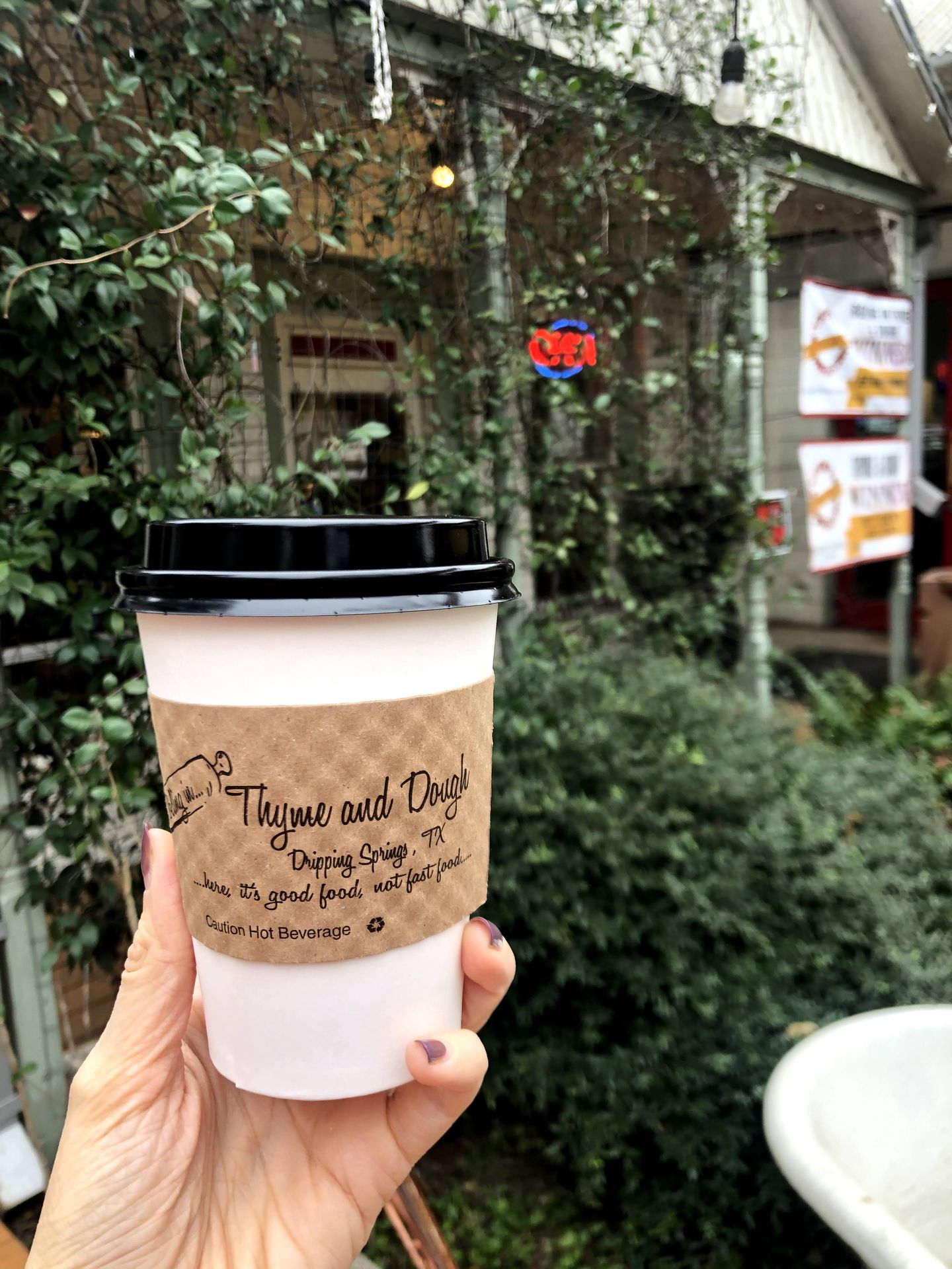 Holding up a coffee in the outdoor patio at Rolling in Thyme and Dough. There is greenery in the background