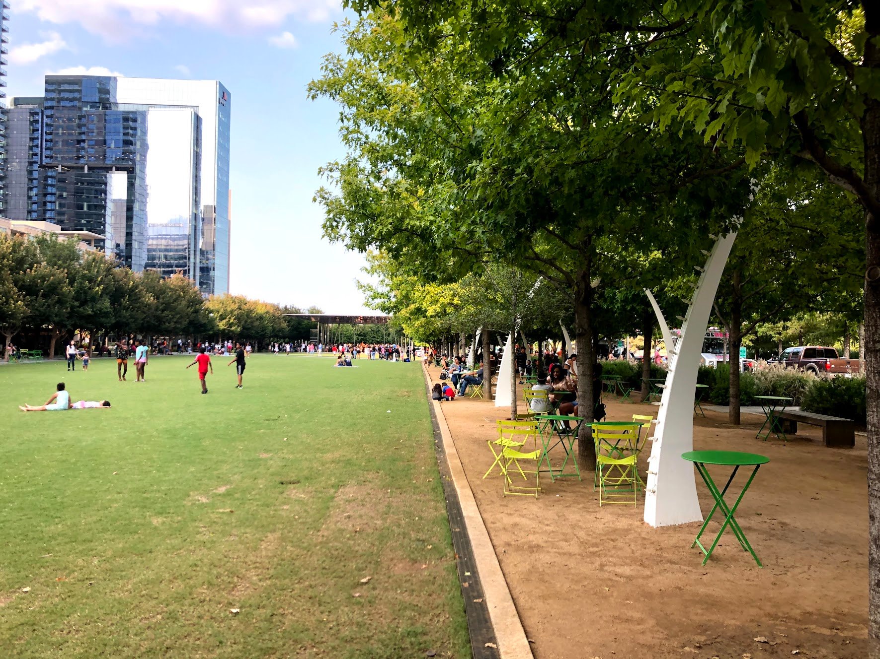 A large lawn and the pathway with white arches with trees. There are tables under the shade of the trees.