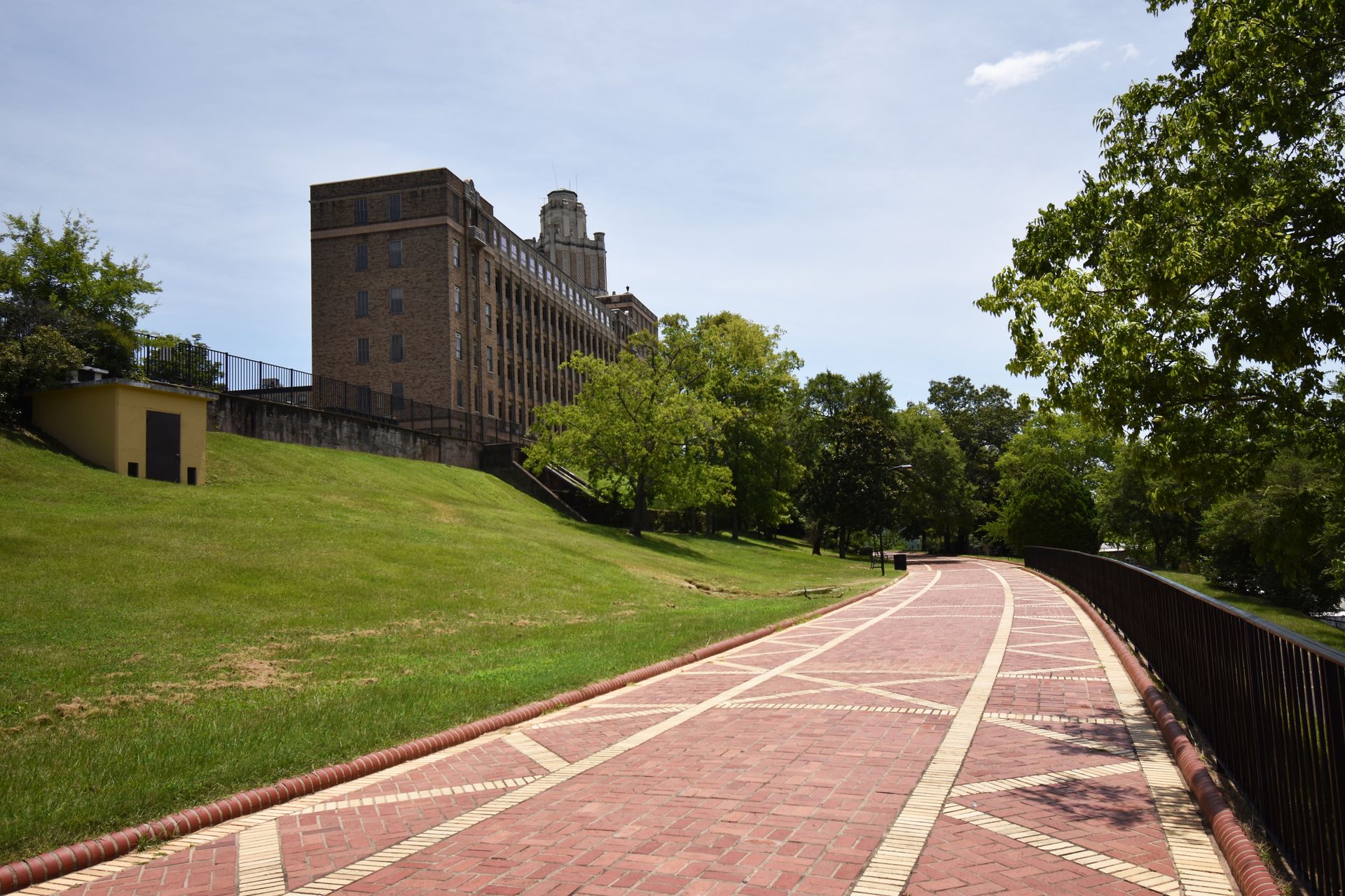 A wide, red and yellow brick promenade. There is a hill with a building on the left and a fence to the right.