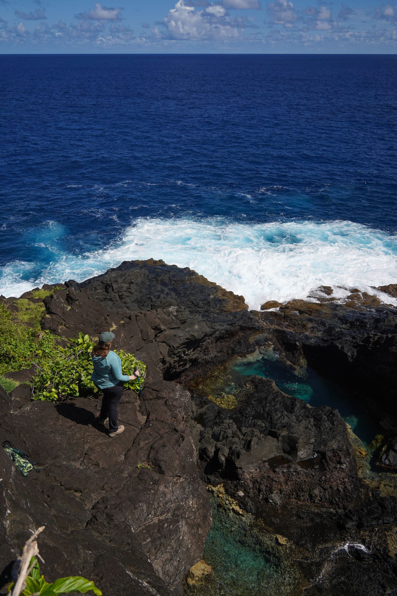 Lydia standing on a black rock coastline with the ocean below her. There are pools of blue water in the black rocks below.