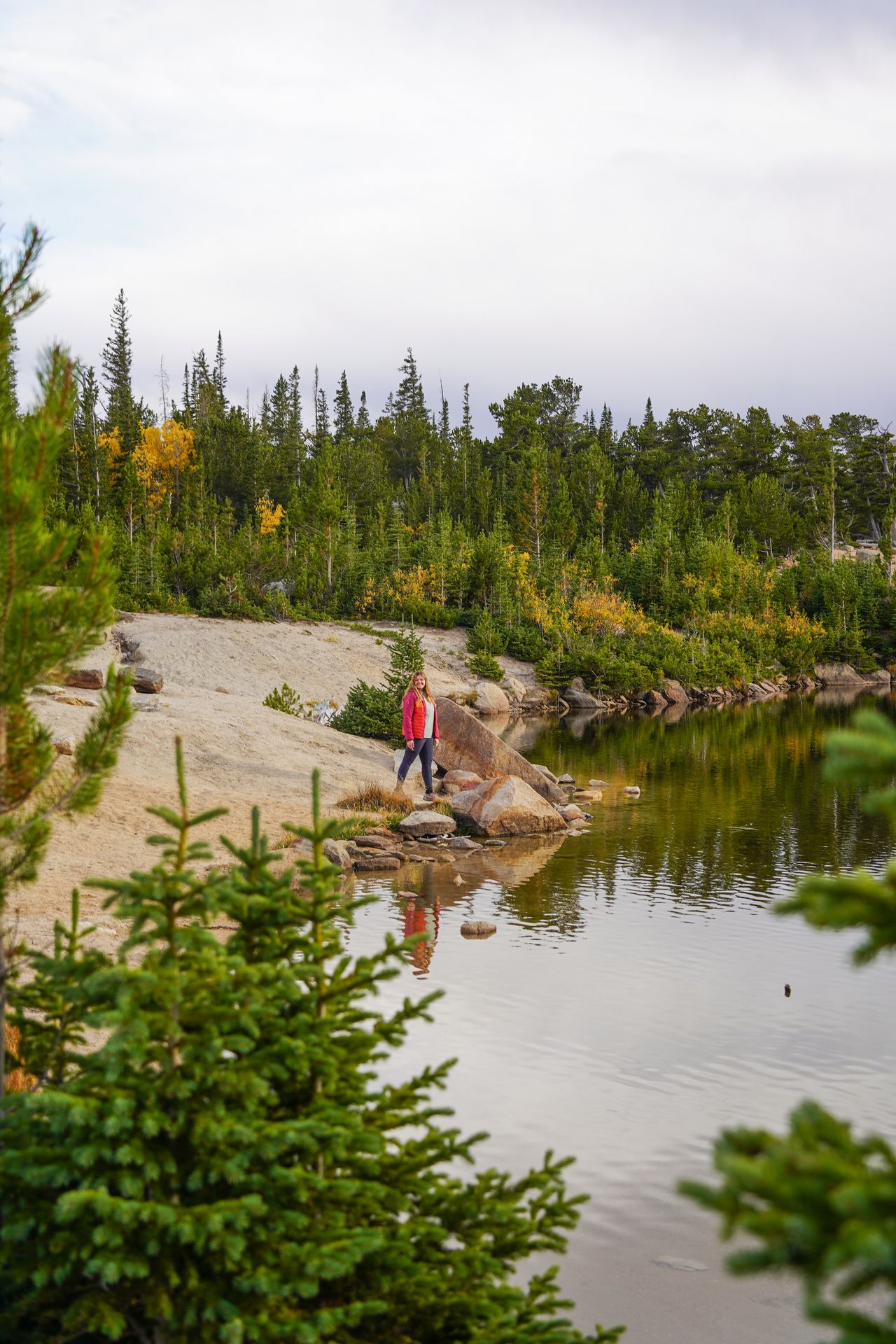 Lydia standing on the shore of a lake that is surrounded by green trees and a bit of yellow foilage