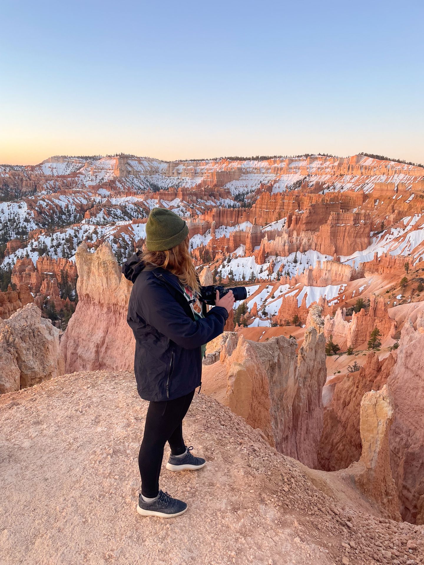 Lydia standing on a rocky cliff and holding a camera while looking at Bryce Canyon with at sunrise