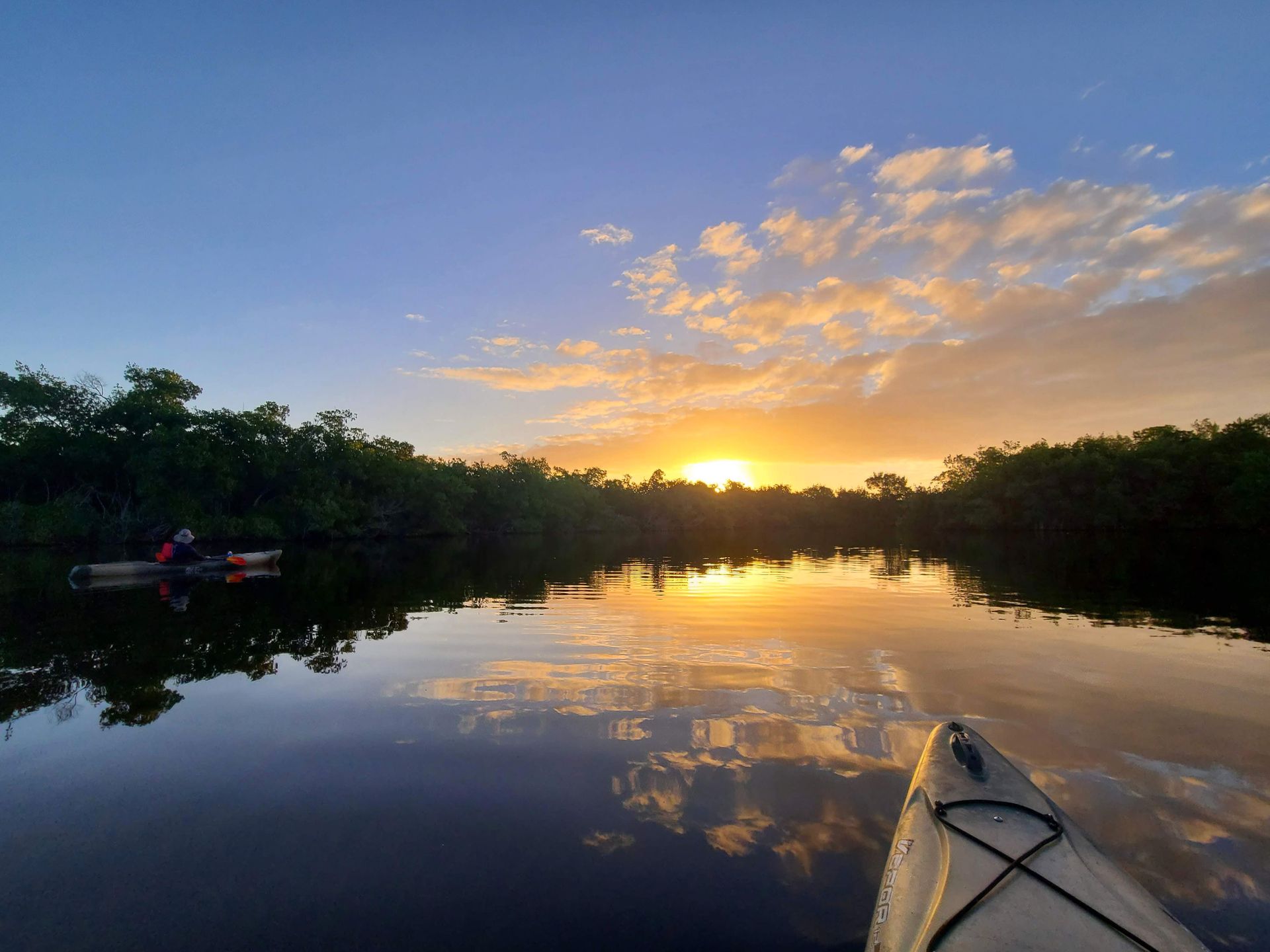 The front of a kayak with a sunrise in the sky and reflecting on the water. Yellow and orange clouds reflect onto the water